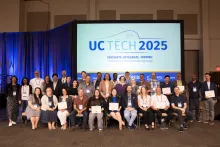 All UC Tech 2025 award winners sit together in three rows and smile for a photo. The UC Tech 2025 logo is displayed on a screen behind the award winners.