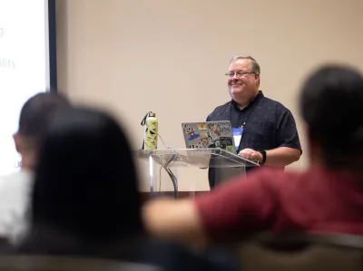 UC Tech 2025 speaker presenting for their session. The backs of several attendees are in the foreground.