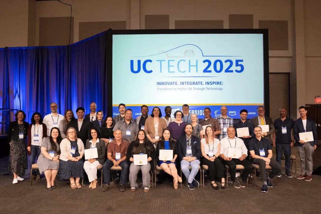 All UC Tech 2025 award winners sit together in three rows and smile for a photo. The UC Tech 2025 logo is displayed on a screen behind the award winners.