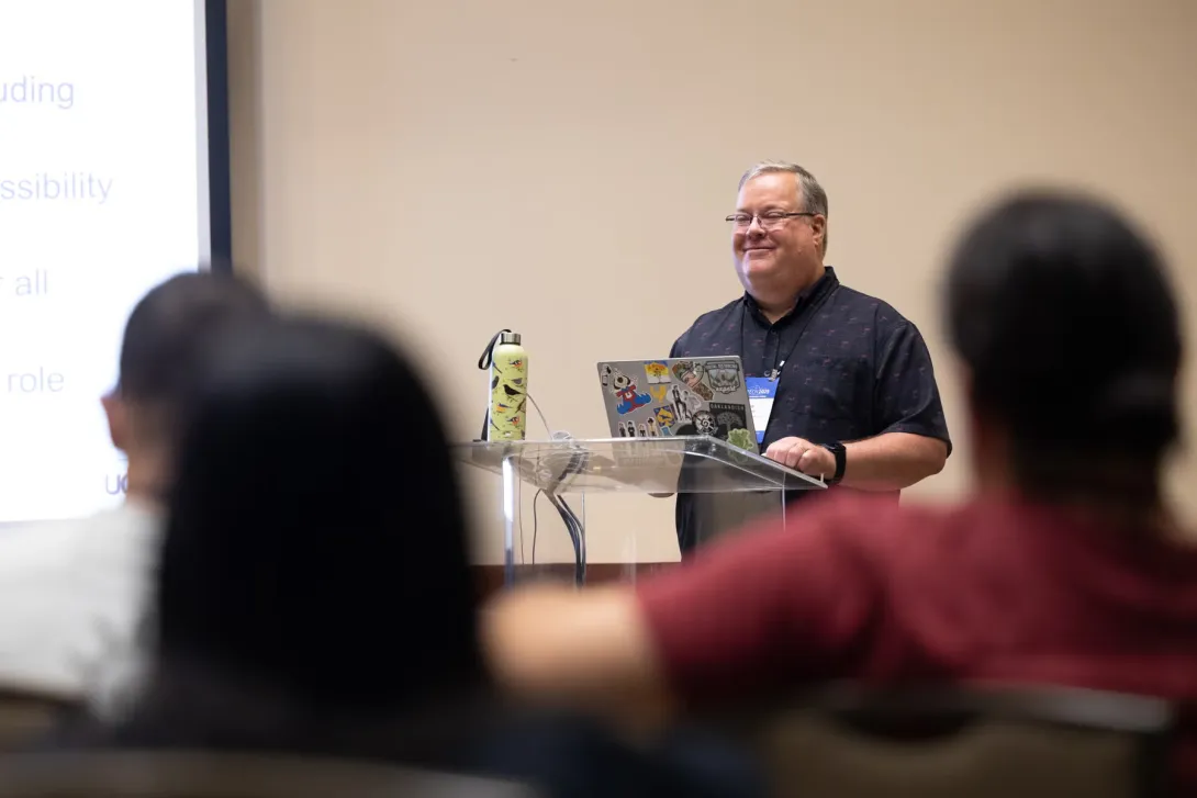 UC Tech 2025 speaker presenting for their session. The backs of several attendees are in the foreground.